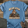 Rear View Included Comfort Colors T-shirt in Blue Jean, laid flat. A cartoon owl in a helmet and goggles rides a muddy ATV past pine trees, looking back over its shoulder, between the words Rear View Included. Olive cargo pants and brown hiking boots lie on a wood floor.