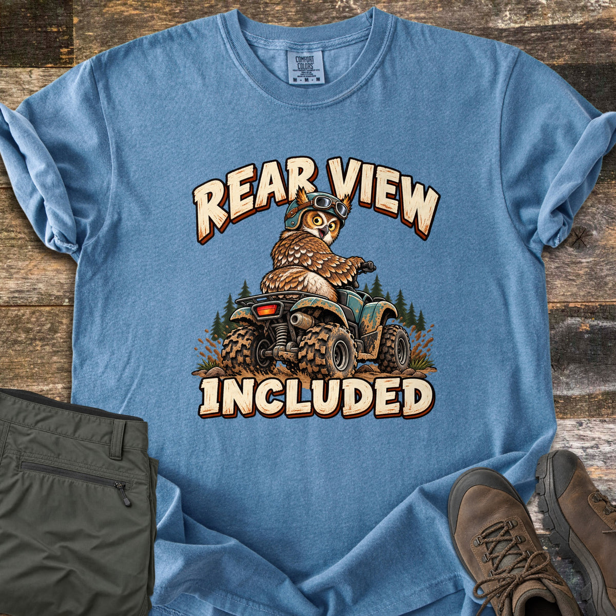Rear View Included Comfort Colors T-shirt in Blue Jean, laid flat. A cartoon owl in a helmet and goggles rides a muddy ATV past pine trees, looking back over its shoulder, between the words Rear View Included. Olive cargo pants and brown hiking boots lie on a wood floor.