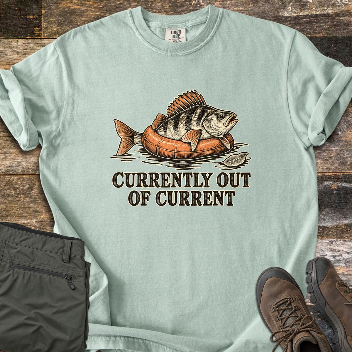 Currently Out of Current Comfort Colors T-shirt in Bay, laid flat. A striped perch lounges on an orange inner tube floating in water beside a leaf, above the words Currently Out of Current. Olive hiking pants and brown hiking boots sit on weathered wood planks.