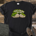You Grow on Me Comfort Colors T-shirt in Black, laid flat. Two smiling cartoon rocks covered in green moss lean together under the words You Grow on Me. Shown on a wood plank background with folded green hiking pants and brown hiking boots.