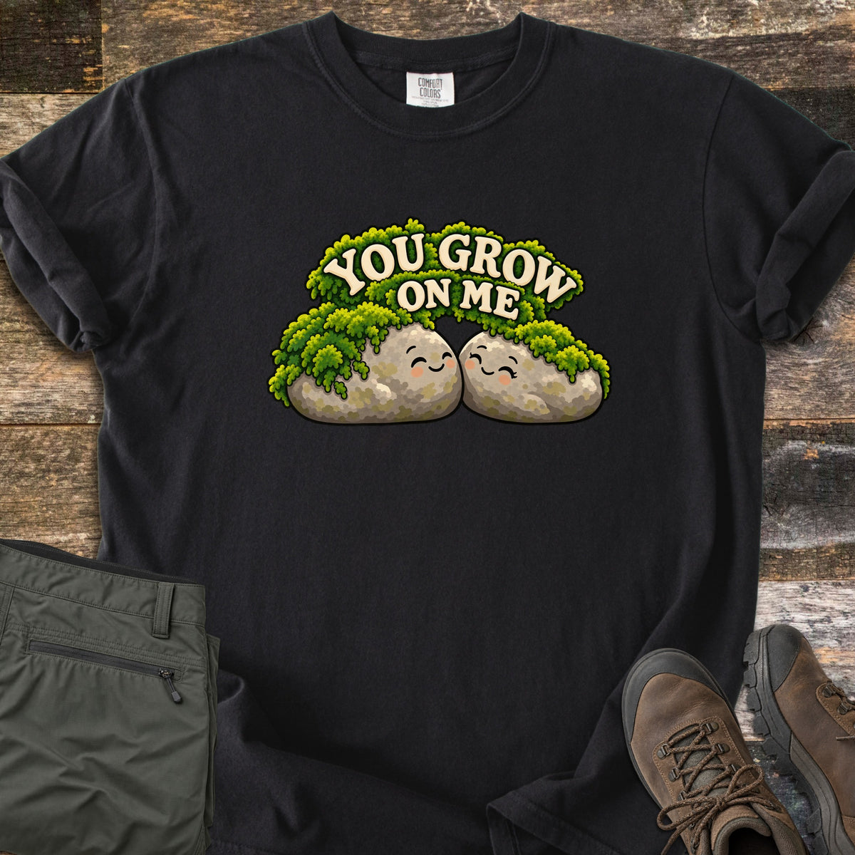 You Grow on Me Comfort Colors T-shirt in Black, laid flat. Two smiling cartoon rocks covered in green moss lean together under the words You Grow on Me. Shown on a wood plank background with folded green hiking pants and brown hiking boots.