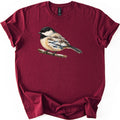 Chickadee Winter Creek Comfort Colors T-shirt in Maroon, laid flat. A painted black-capped chickadee perches on a snowy branch, its body filled with a winter scene of a creek, snowbanks and bare trees. No words are printed. The shirt lies on a white surface with rolled sleeves.