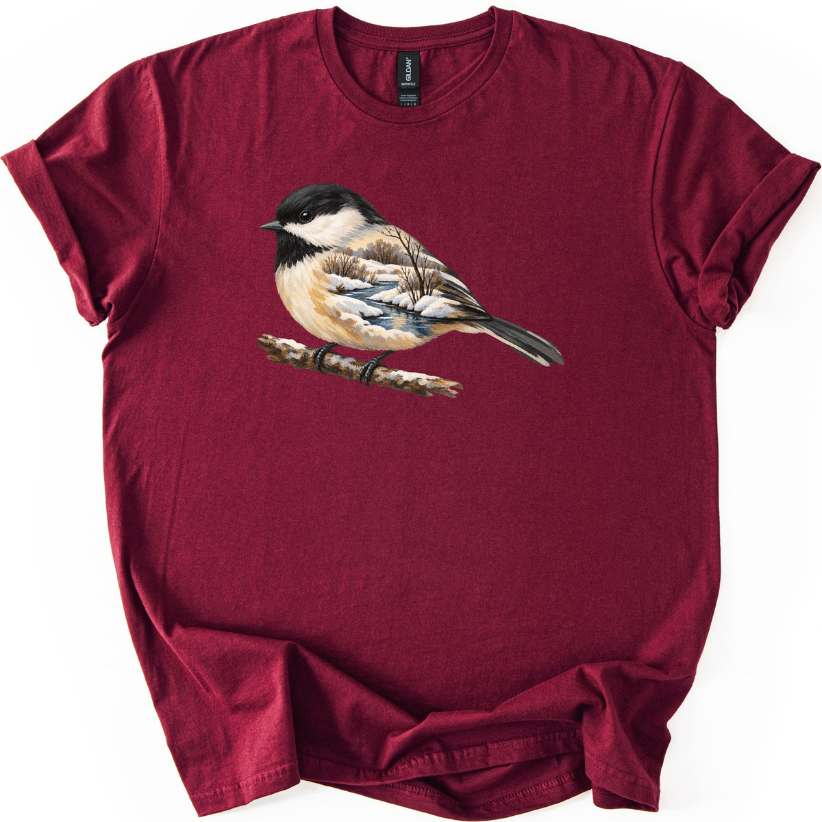 Chickadee Winter Creek Comfort Colors T-shirt in Maroon, laid flat. A painted black-capped chickadee perches on a snowy branch, its body filled with a winter scene of a creek, snowbanks and bare trees. No words are printed. The shirt lies on a white surface with rolled sleeves.