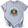 Life Is Better By The Fire Badge Comfort Colors T-shirt in Sport Grey, laid flat. A circular badge shows a campfire of crossed logs in front of green pine trees, with LIFE IS BETTER arched above in cream and BY THE FIRE below in dark green. Sleeves are cuffed on a white background.