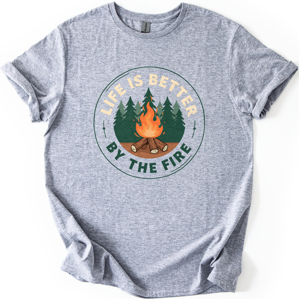 Life Is Better By The Fire Badge Comfort Colors T-shirt in Sport Grey, laid flat. A circular badge shows a campfire of crossed logs in front of green pine trees, with LIFE IS BETTER arched above in cream and BY THE FIRE below in dark green. Sleeves are cuffed on a white background.