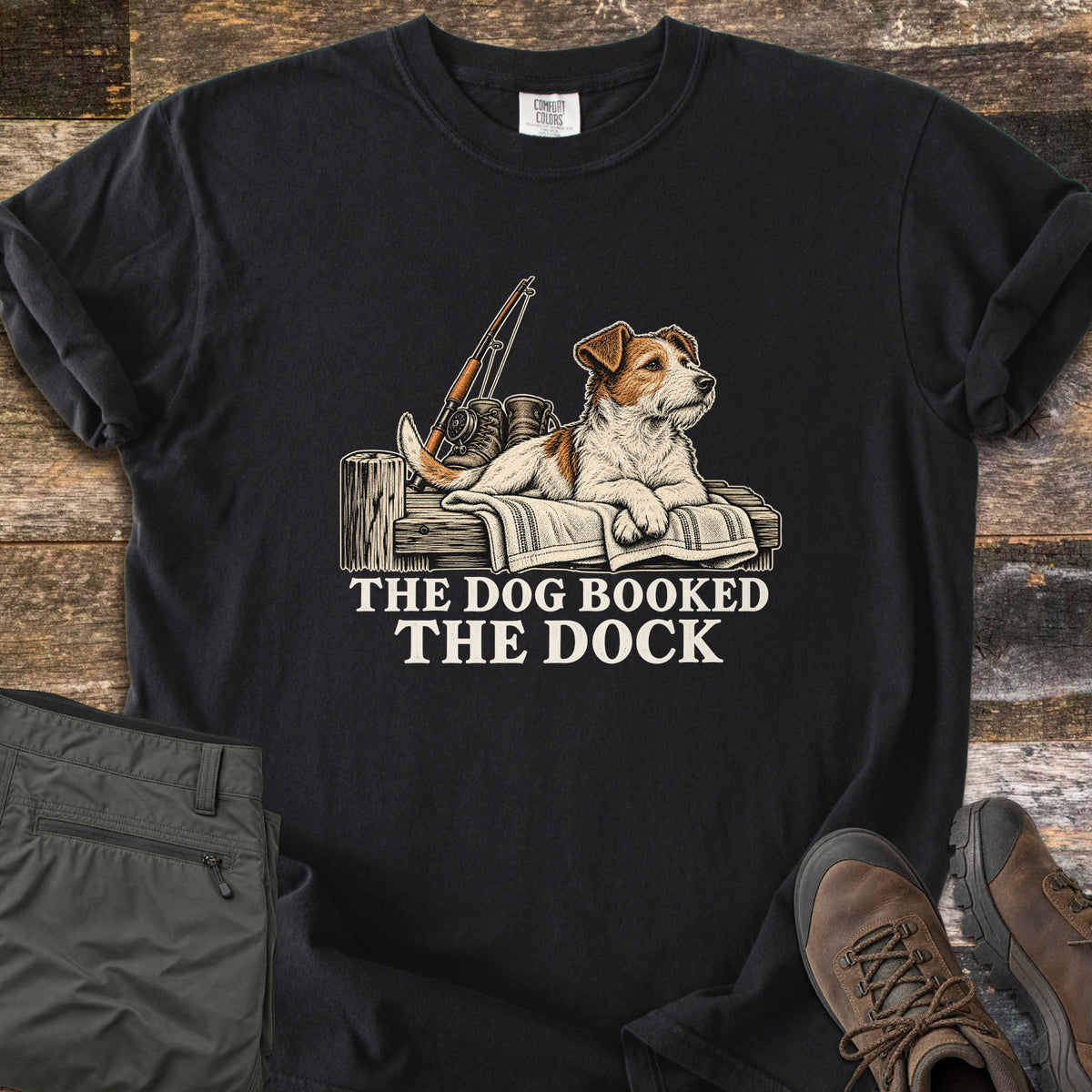 The Dog Booked the Dock Comfort Colors T-shirt in Black, laid flat. An illustrated Jack Russell terrier lies on a towel on a wooden dock beside a fishing rod and boots, above the words The Dog Booked The Dock. Olive pants and brown hiking boots sit on weathered wood.