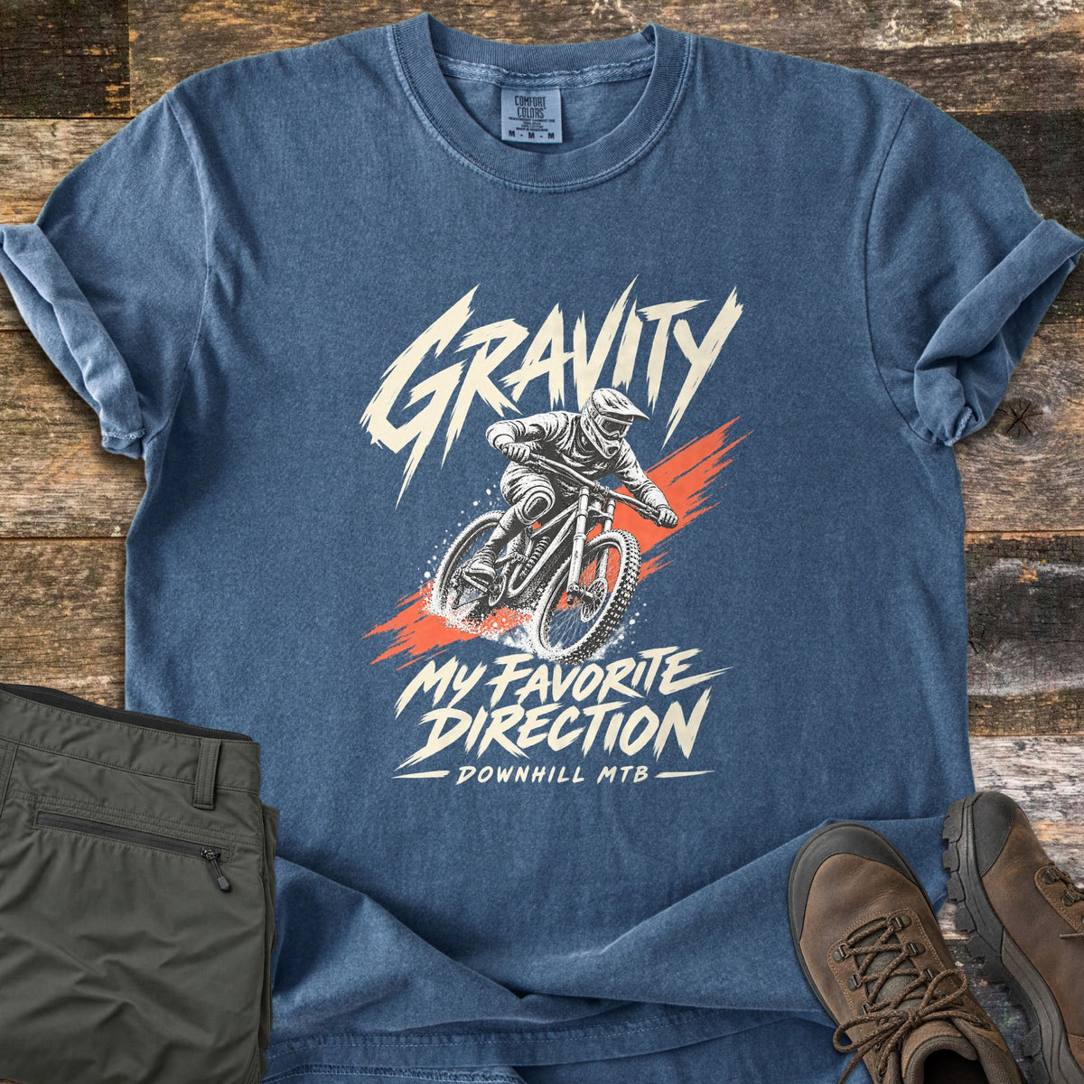 Gravity My Favorite Direction Comfort Colors T-shirt in Denim, laid flat. A downhill mountain biker in a full-face helmet kicks up dirt over an orange brush stroke, with Gravity above and My Favorite Direction, Downhill MTB below. Olive pants and brown hiking boots sit on weathered wood.