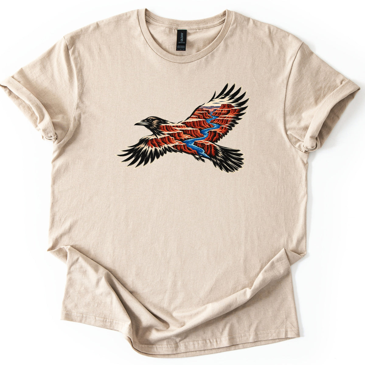 Red Canyon Raven Comfort Colors T-shirt in Sand, laid flat. A black raven in flight, its body and wings filled with red canyon cliffs and a winding blue river. No words are printed. The shirt lies on a white surface with rolled sleeves and a loosely folded hem.