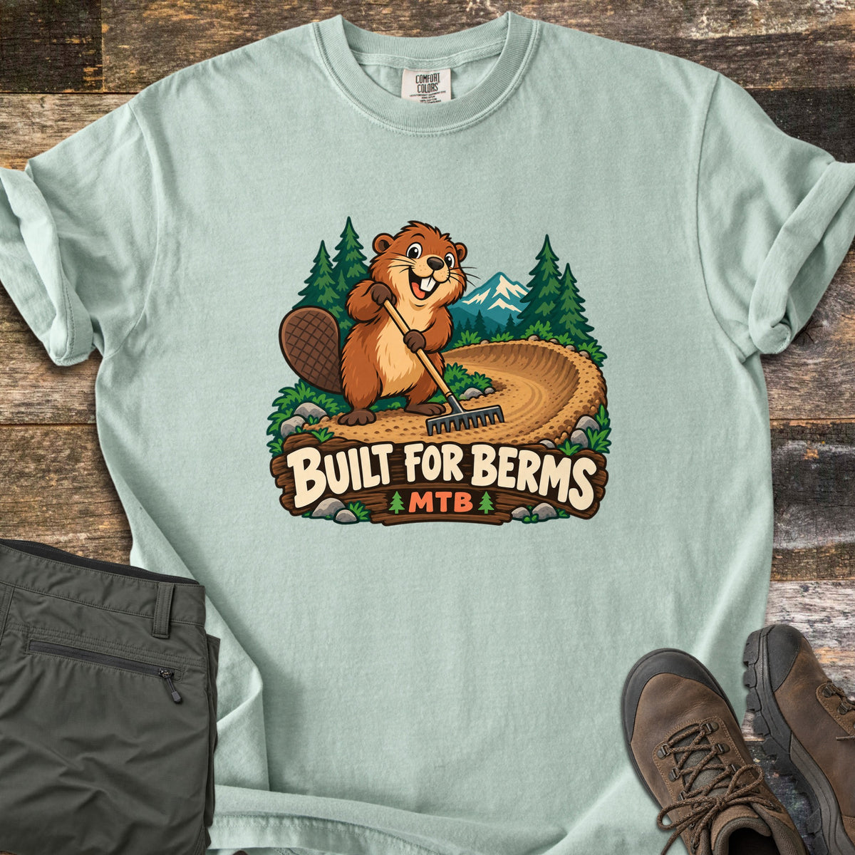 Built For Berms Comfort Colors T-shirt in Bay, laid flat. A cartoon beaver holds a rake on a curved dirt berm, with pine trees and a snowy mountain behind, above a wooden sign reading Built For Berms MTB. Olive pants and brown hiking boots lie beside it on weathered wood.
