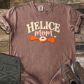 Helice Mom Comfort Colors T-shirt in Espresso, laid flat. HELICE in cream letters over mom in pink script with a small heart, above an orange two-winged helice target with a white center. Folded olive pants and brown hiking boots lie beside it on weathered wood.