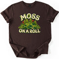Moss on a Roll Comfort Colors T-shirt in Dark Chocolate, laid flat. Cream lettering reads MOSS above and ON A ROLL below a mound of green paper-quilled moss swirls and fronds with two rust-brown spore capsules. The shirt has cuffed sleeves on a plain white background.