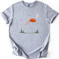 Moonrise Clay Comfort Colors T-shirt in Sport Grey, laid flat. An orange clay target streaks across a night sky with a cream crescent moon and small green stars, above pine trees and low hills. No words are printed. Sleeves rolled, on a white background.