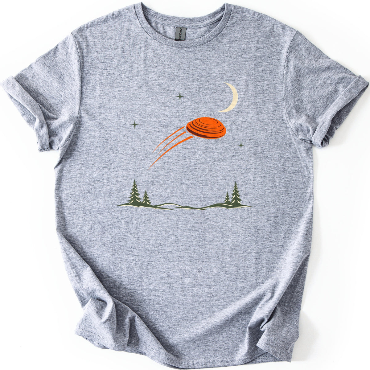 Moonrise Clay Comfort Colors T-shirt in Sport Grey, laid flat. An orange clay target streaks across a night sky with a cream crescent moon and small green stars, above pine trees and low hills. No words are printed. Sleeves rolled, on a white background.