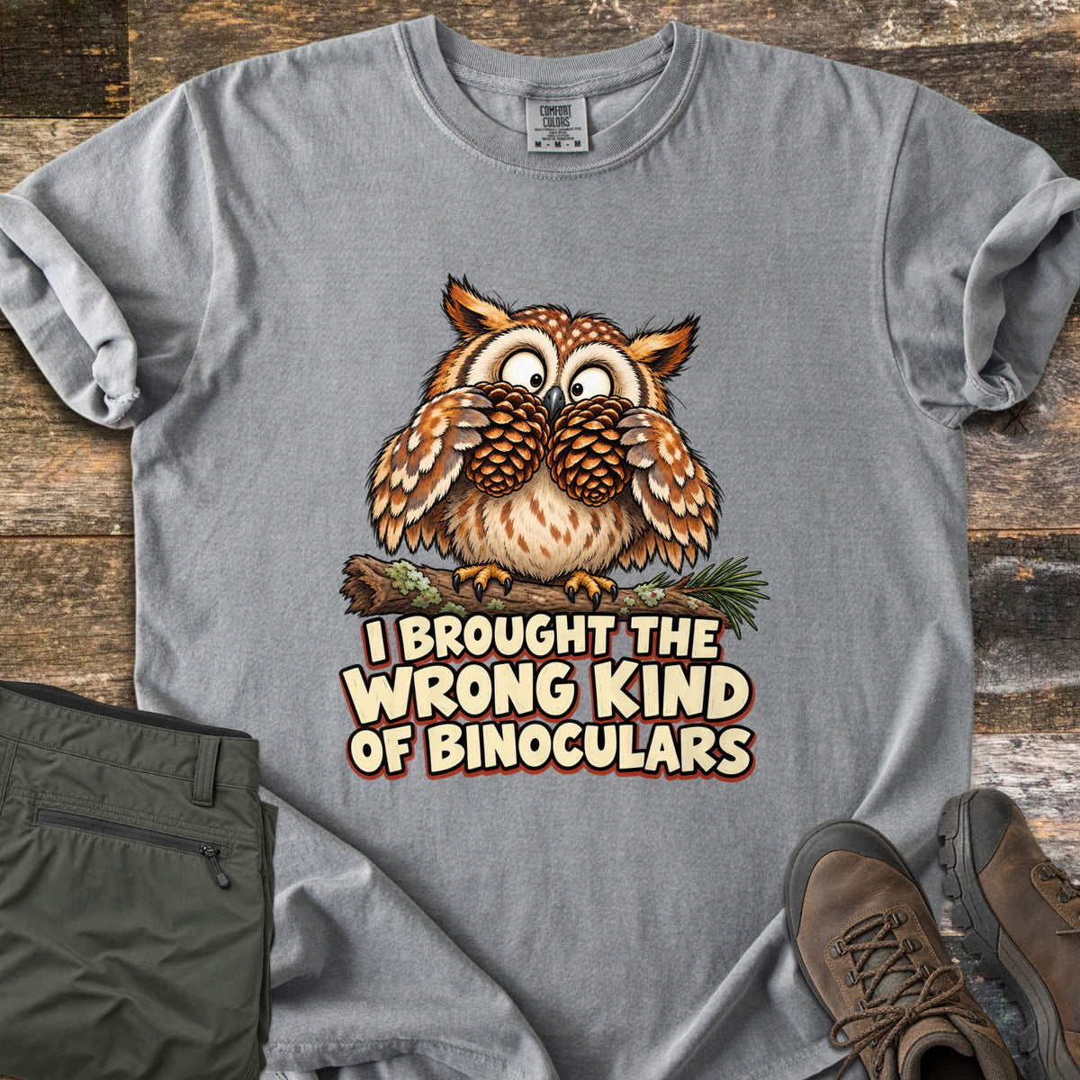 I Brought The Wrong Kind Of Binoculars Comfort Colors T-shirt in Grey, laid flat. A wide-eyed cartoon owl on a mossy branch holds two pinecones to its eyes like binoculars above the words I Brought The Wrong Kind Of Binoculars. Olive hiking pants and brown hiking boots lie beside it on weathered wood.