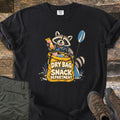 Snack Department Comfort Colors T-shirt in Black, laid flat. A raccoon in a blue life vest holds a snack bar over a yellow dry bag full of snacks, a paddle beside it, with the words Dry Bag Snack Department. Olive pants and brown hiking boots sit on wood planks.