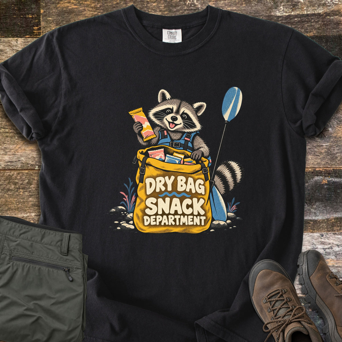 Snack Department Comfort Colors T-shirt in Black, laid flat. A raccoon in a blue life vest holds a snack bar over a yellow dry bag full of snacks, a paddle beside it, with the words Dry Bag Snack Department. Olive pants and brown hiking boots sit on wood planks.