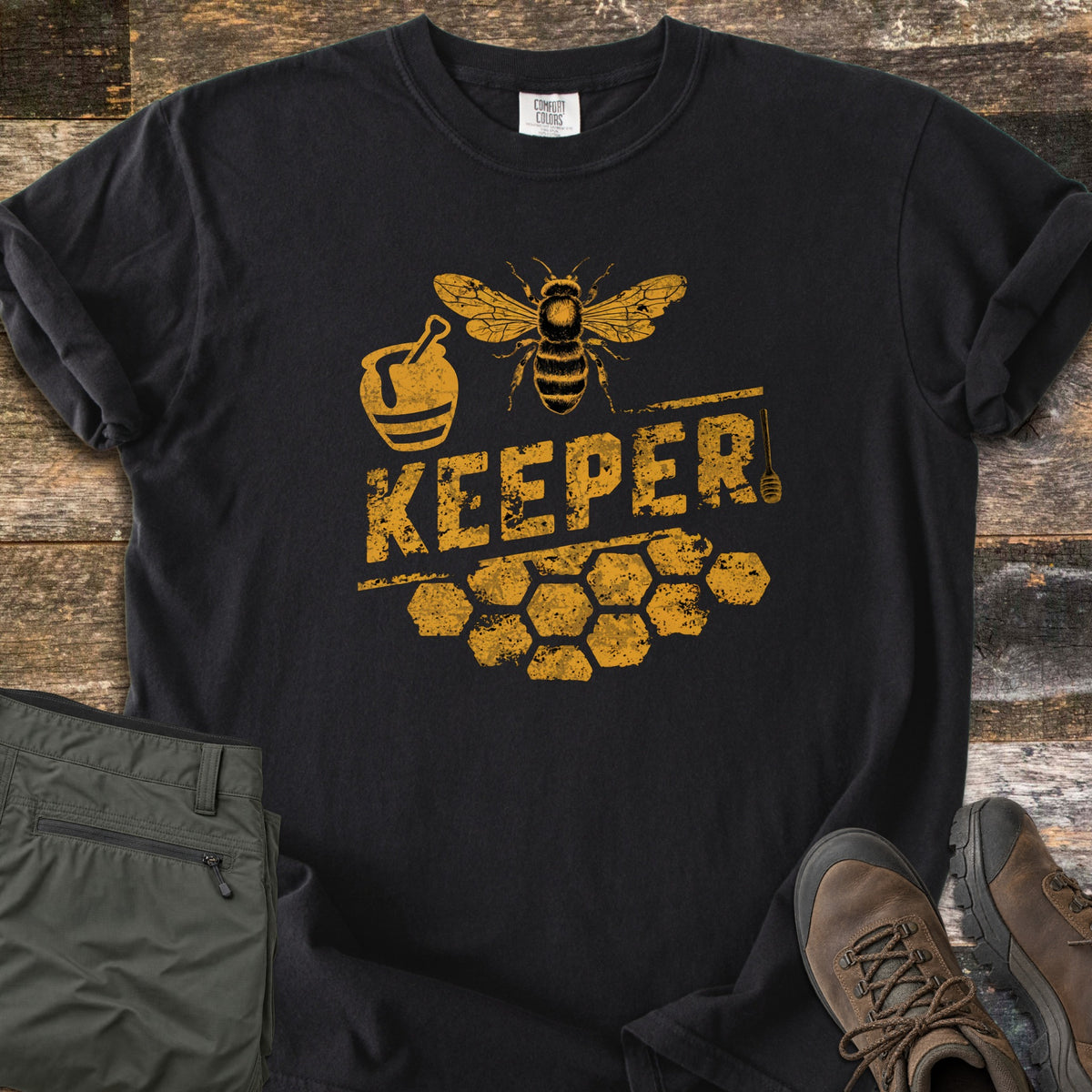 Bee Keeper Comfort Colors T-shirt in Black, laid flat. Distressed gold print of a bee above the word KEEPER, with a honey pot, a honey dipper and honeycomb cells. The shirt lies on weathered wood beside folded green pants and brown hiking boots.