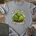 My Social Circle Is a Terrarium Comfort Colors T-shirt in Grey, laid flat. A glass bowl terrarium holds three smiling moss balls among rocks and soil, between the words My Social Circle and Is a Terrarium. Folded olive pants and brown hiking boots sit on weathered wood planks.