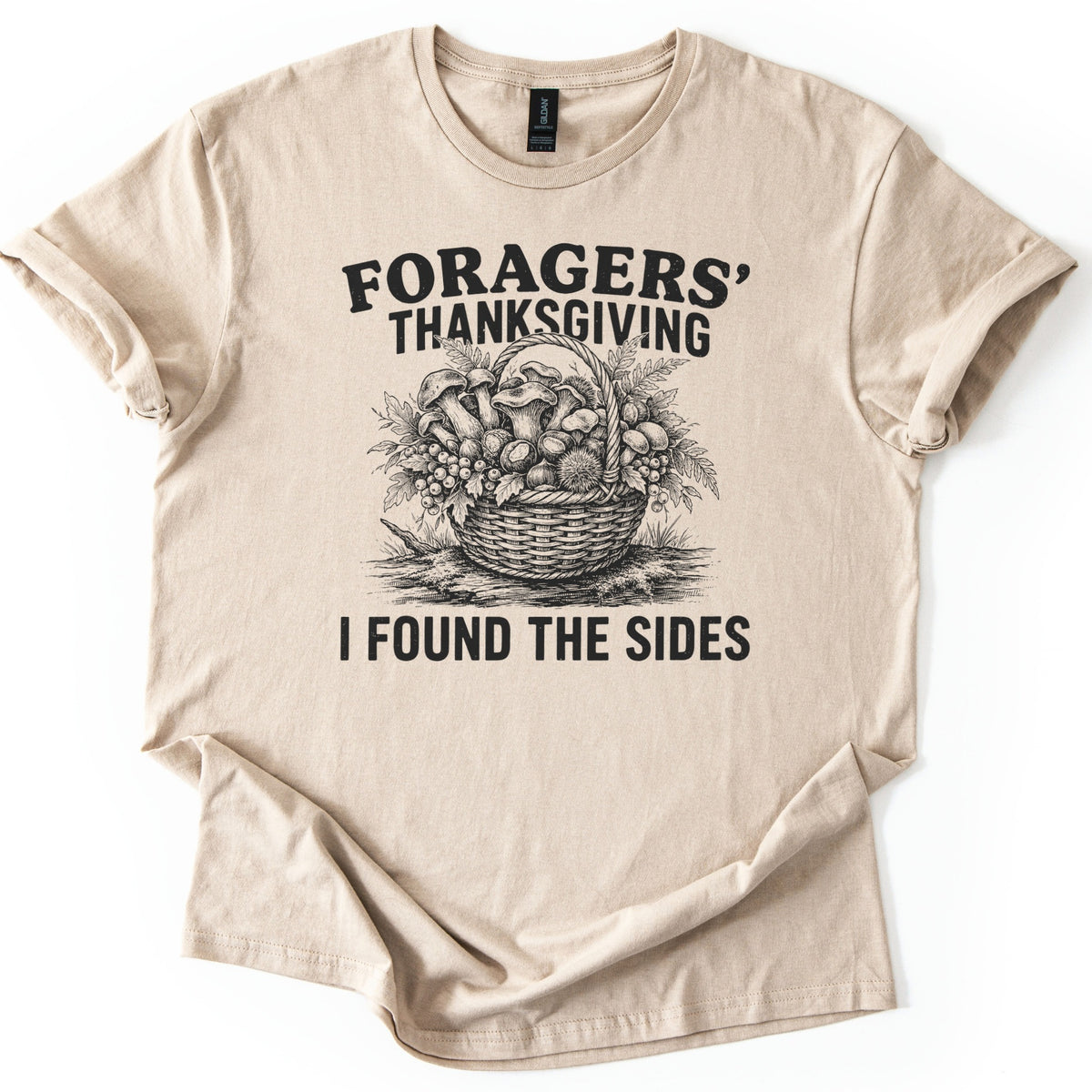 Foragers Thanksgiving Comfort Colors T-shirt in Sand, laid flat. Black sketch of a wicker basket of mushrooms, berries, ferns and a chestnut burr, between the words Foragers' Thanksgiving and I Found the Sides. Sleeves cuffed, on a white background.