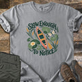 Slow Enough To Notice Comfort Colors T-shirt in Grey, laid flat. A coral canoe with a yellow paddle floats among lily pads, a water lily, cattails and a dragonfly between the words Slow Enough To Notice. Folded green hiking pants and brown hiking boots sit on weathered wood.