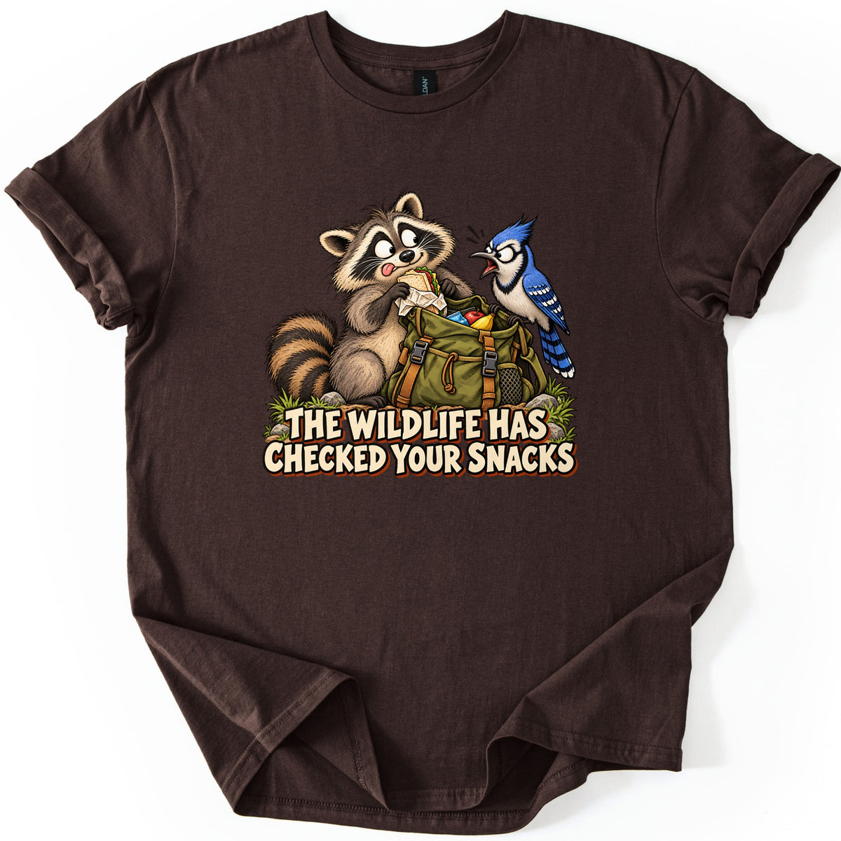 The Wildlife Has Checked Your Snacks Comfort Colors T-shirt in Dark Chocolate, laid flat. A cartoon raccoon eats a sandwich from an open green backpack while a blue jay squawks at it, above the words The Wildlife Has Checked Your Snacks. The shirt has rolled sleeves on a white background.