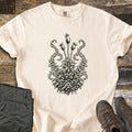 Moss Woodcut Comfort Colors T-shirt in Ivory, laid flat. A black woodcut-style print of a moss clump with three tall spore capsules on stems and curling fronds on each side. Olive cargo pants and brown hiking boots sit beside it on weathered wood planks.
