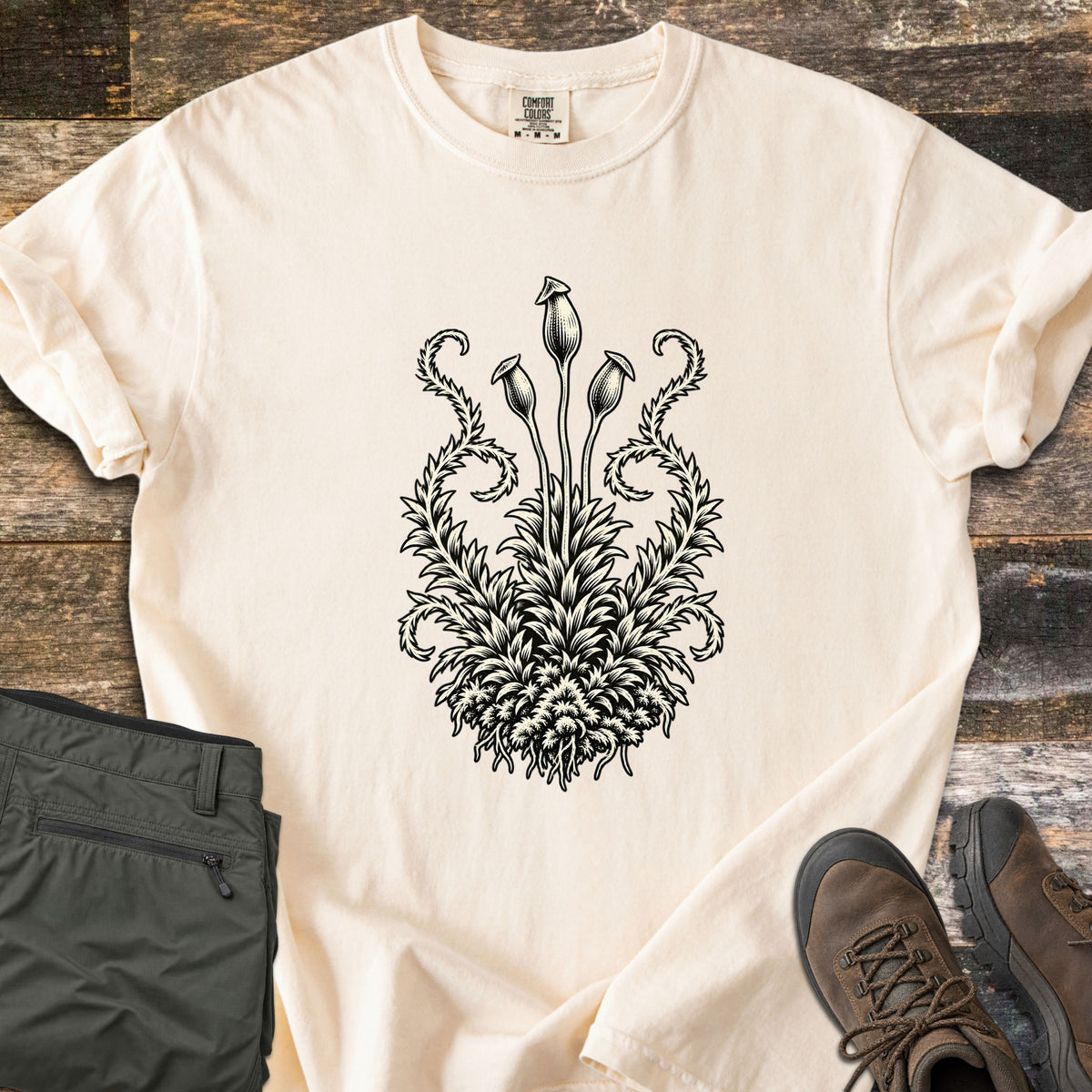 Moss Woodcut Comfort Colors T-shirt in Ivory, laid flat. A black woodcut-style print of a moss clump with three tall spore capsules on stems and curling fronds on each side. Olive cargo pants and brown hiking boots sit beside it on weathered wood planks.