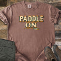 Paddle On Comfort Colors T-shirt in Espresso, laid flat. The words Paddle On in distressed cream lettering with wood-grain edges and rivets, above a wooden canoe paddle. Folded green hiking pants and brown hiking boots sit on a weathered wood floor.