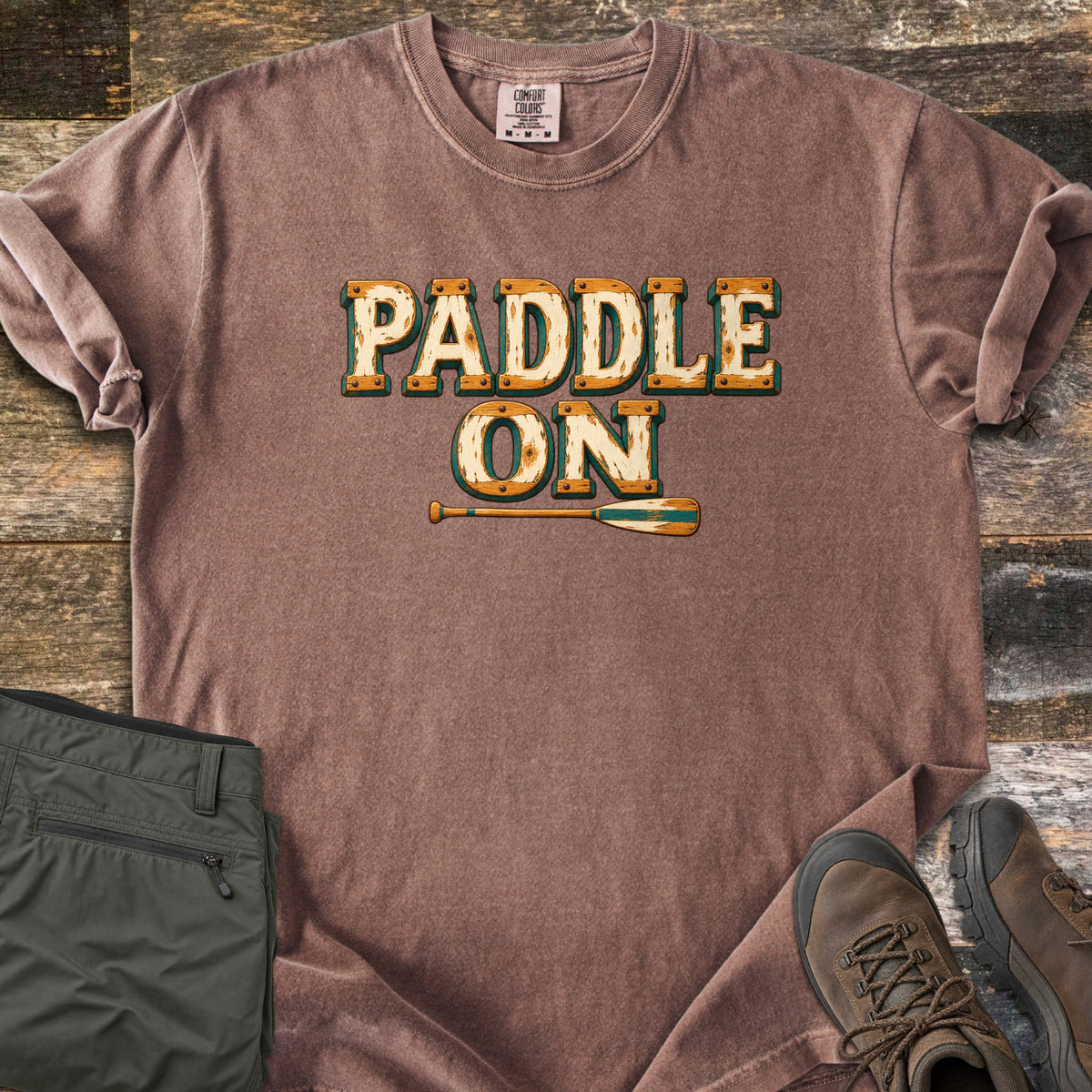 Paddle On Comfort Colors T-shirt in Espresso, laid flat. The words Paddle On in distressed cream lettering with wood-grain edges and rivets, above a wooden canoe paddle. Folded green hiking pants and brown hiking boots sit on a weathered wood floor.