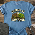 Dormant Not Done Comfort Colors T-shirt in Blue Jean, laid flat. Cream letters read Dormant over a mound of moss on a rock, dry brown on the left and green on the right, with a water drop above, and Not Done below. Olive pants and brown hiking boots lie on weathered wood.