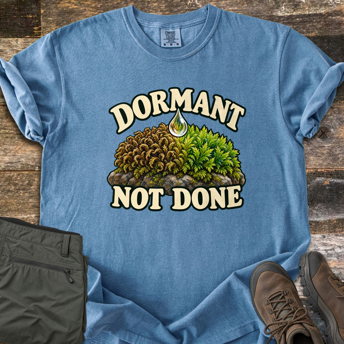 Dormant Not Done Comfort Colors T-shirt in Blue Jean, laid flat. Cream letters read Dormant over a mound of moss on a rock, dry brown on the left and green on the right, with a water drop above, and Not Done below. Olive pants and brown hiking boots lie on weathered wood.