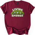 Living Sponge Comfort Colors T-shirt in Maroon, laid flat. A mound of bright green moss on gray rocks holding three clear water droplets, with LIVING arched above and SPONGE below in cream bubble letters. The shirt has cuffed sleeves on a white background.
