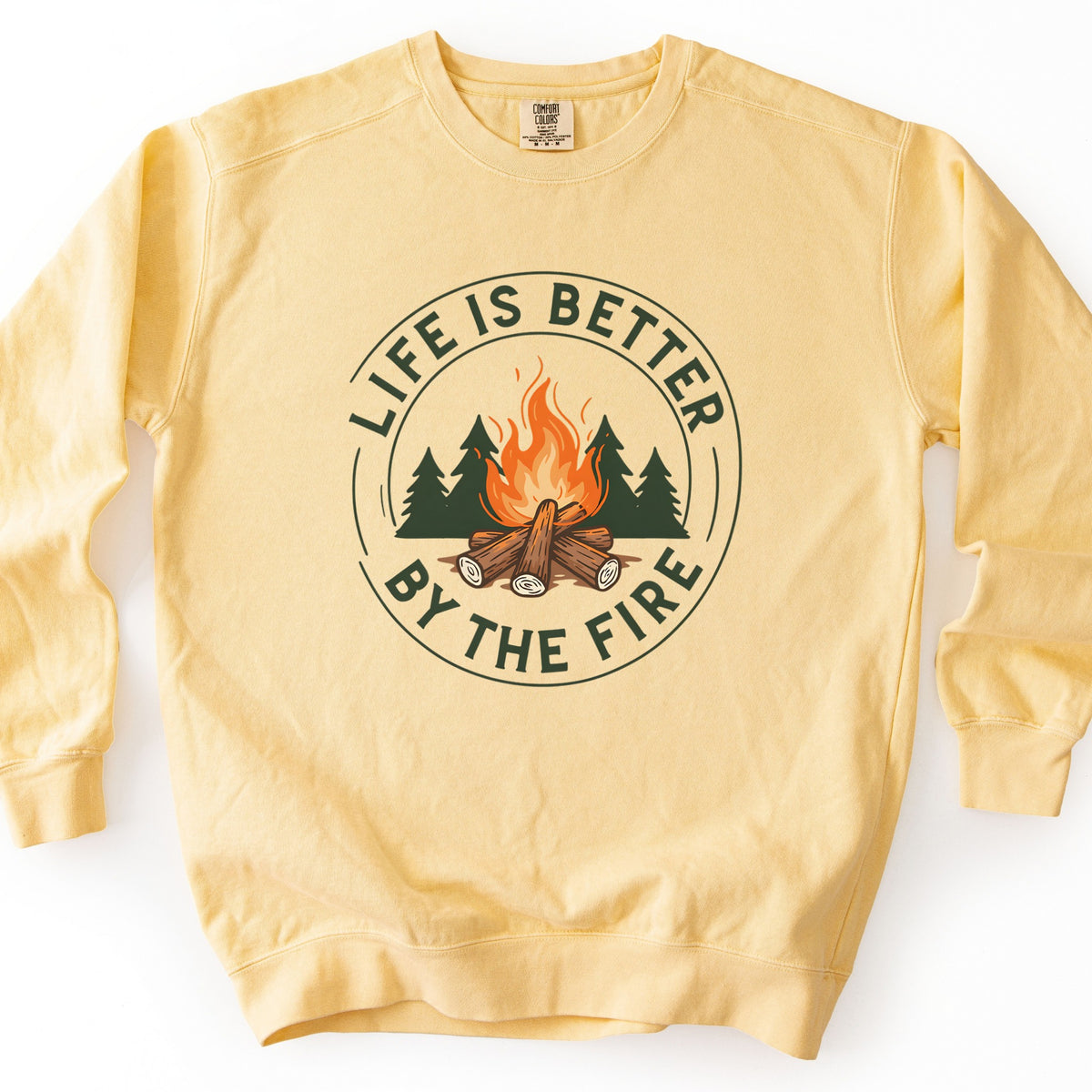 Life Is Better By The Fire Ring Comfort Colors crewneck sweatshirt in Butter, laid flat. A circular badge shows a campfire of crossed logs with orange flames in front of dark green pine trees, ringed by the words Life Is Better By The Fire in dark green. It lies on a white surface.