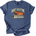 Dirt Sculptor Comfort Colors T-shirt in Heather Navy, laid flat. Dirt Sculptor arches over a curved dirt berm on a stone pedestal reading My Gallery Has Berms, MTB Trail Crew, with a shovel leaning against it. The shirt has rolled sleeves on a white background.
