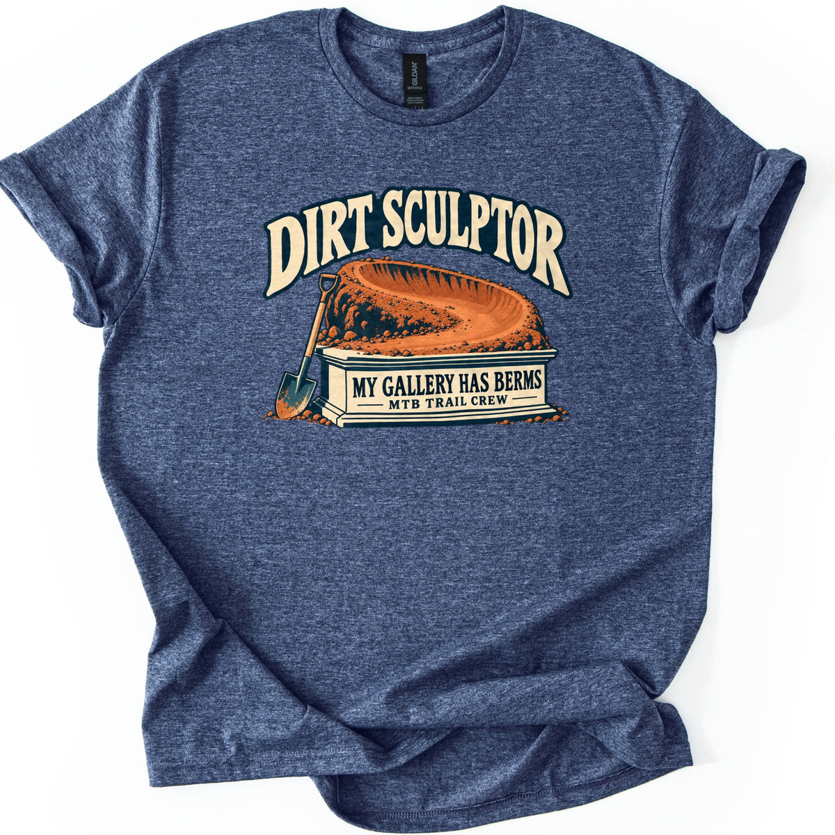 Dirt Sculptor Comfort Colors T-shirt in Heather Navy, laid flat. Dirt Sculptor arches over a curved dirt berm on a stone pedestal reading My Gallery Has Berms, MTB Trail Crew, with a shovel leaning against it. The shirt has rolled sleeves on a white background.