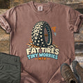 Fat Tires Tiny Worries Comfort Colors T-shirt in Espresso, laid flat. A knobby fat bike tire kicking up dirt, with orange and blue squiggles, above the words Fat Tires Tiny Worries MTB. Sleeves rolled, on weathered wood with olive pants and brown hiking boots.