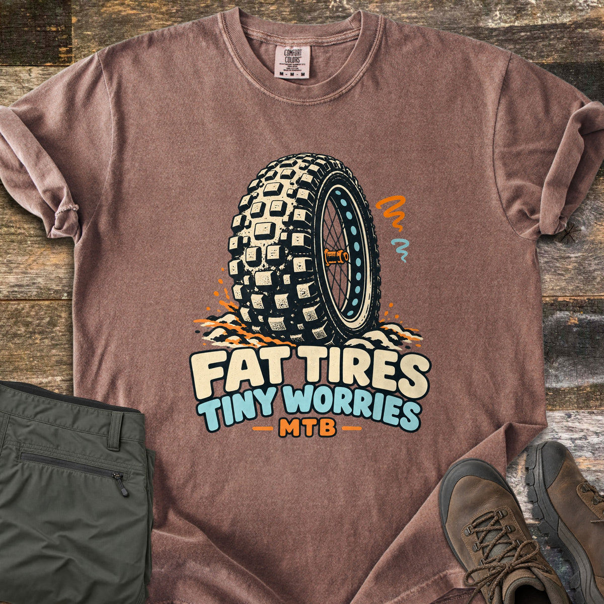 Fat Tires Tiny Worries Comfort Colors T-shirt in Espresso, laid flat. A knobby fat bike tire kicking up dirt, with orange and blue squiggles, above the words Fat Tires Tiny Worries MTB. Sleeves rolled, on weathered wood with olive pants and brown hiking boots.