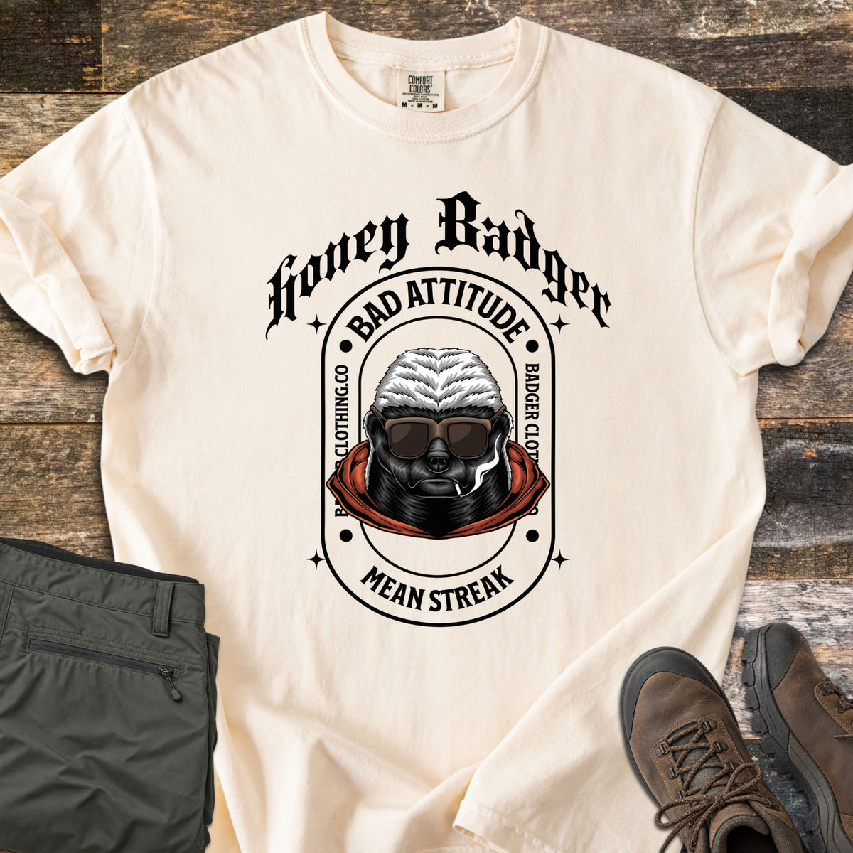 Bad Vibes Badger Comfort Colors T-shirt in Ivory, laid flat. A honey badger in sunglasses and a red hood smokes a cigarette in an arch reading Bad Attitude, Badger Clothing.co, Mean Streak, under Honey Badger in black gothic text. Olive pants and brown boots lie on wood.