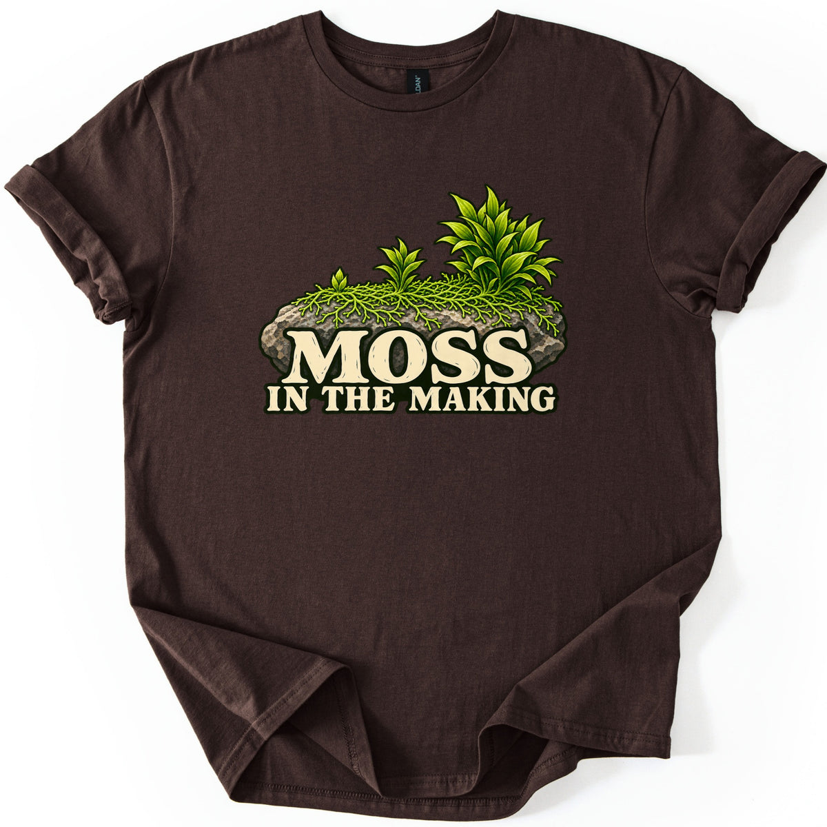 Moss in the Making Comfort Colors T-shirt in Dark Chocolate, laid flat. A mossy gray rock topped with leafy green plants sits above the words MOSS IN THE MAKING in cream lettering. The sleeves are rolled and the shirt lies on a white background.