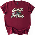 Gone Drifting Comfort Colors T-shirt in Maroon, laid flat. Cream brush-style letters read Gone Drifting around a helmeted rider in an orange jacket on a teal and white snowmobile with a swirling teal trail. The shirt lies on a white background with cuffed sleeves.