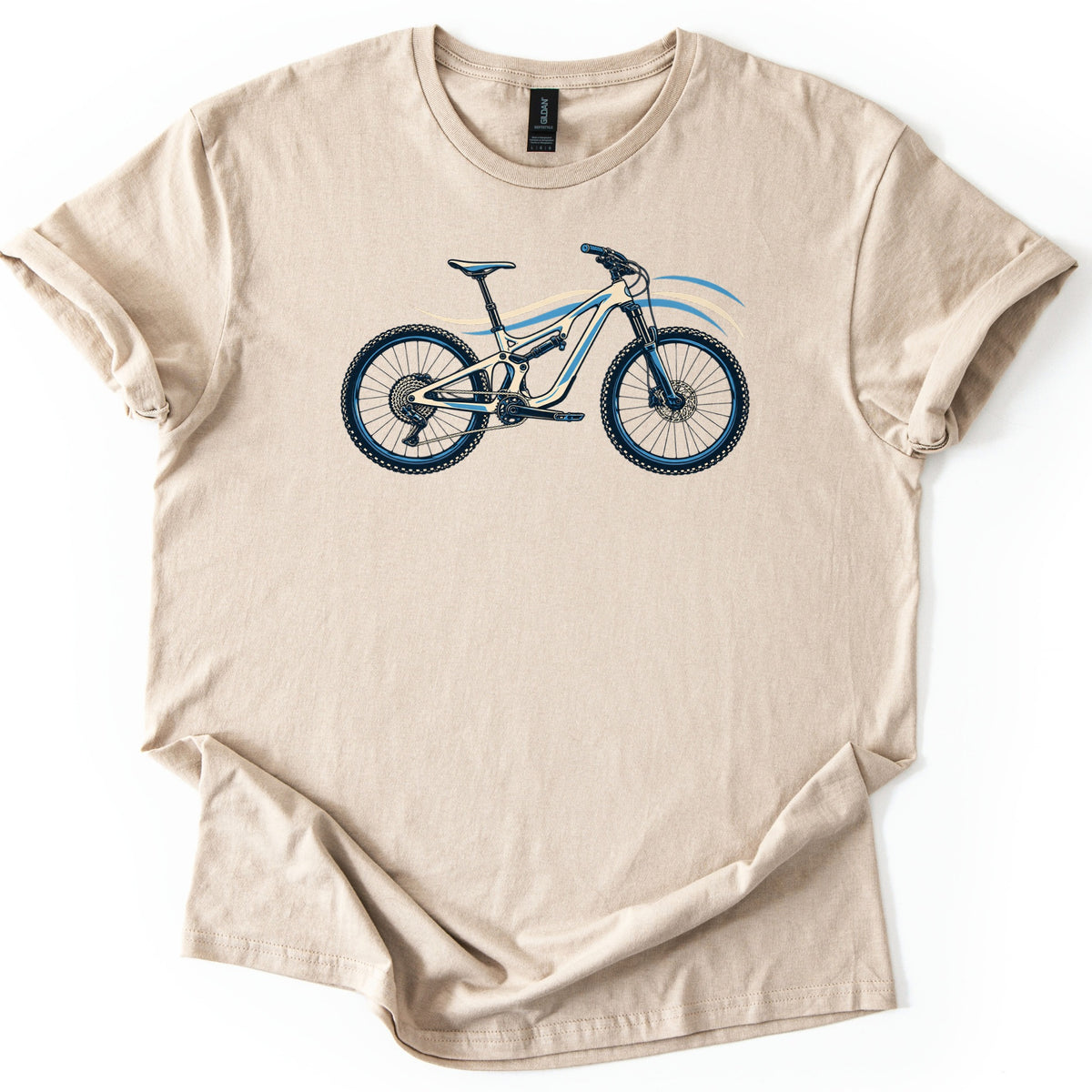 Full Suspension Mountain Bike Comfort Colors T-shirt in Sand, laid flat. A cream and blue full suspension mountain bike with knobby tires and disc brakes is printed across the chest, with flowing blue and cream lines behind it. No words. The sleeves are rolled on a white background.