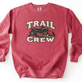 Trail Crew Comfort Colors crewneck sweatshirt in Crimson, laid flat. The words Trail Crew in cream block letters frame a black ATV parked over a fallen branch, with an orange bow saw laid across the front. The sweatshirt lies on a plain white background.