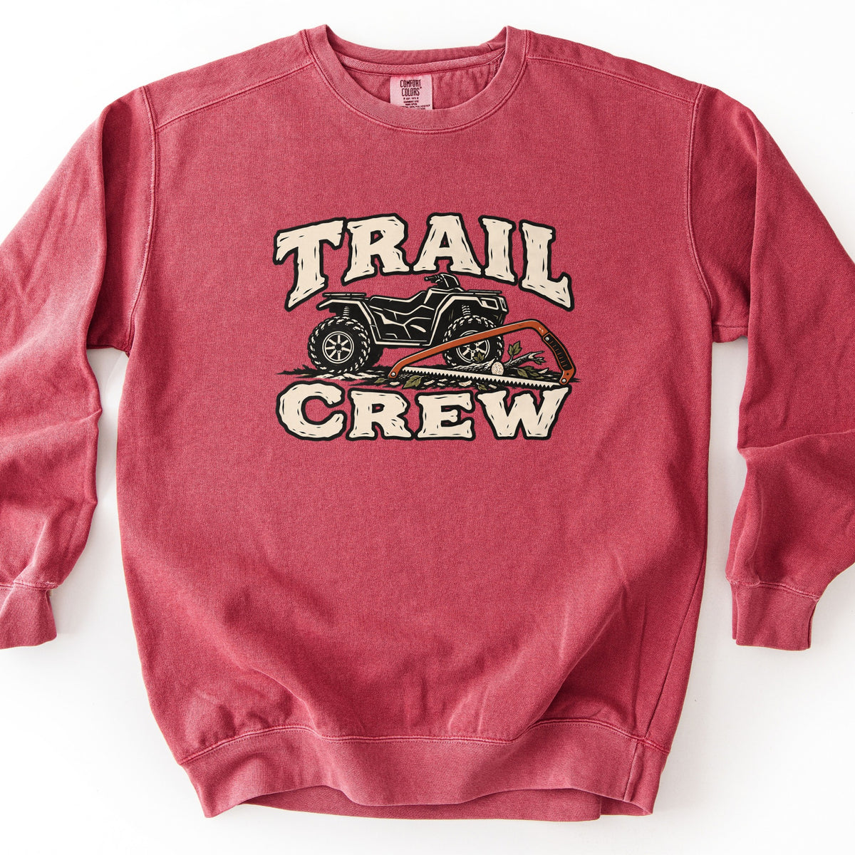 Trail Crew Comfort Colors crewneck sweatshirt in Crimson, laid flat. The words Trail Crew in cream block letters frame a black ATV parked over a fallen branch, with an orange bow saw laid across the front. The sweatshirt lies on a plain white background.