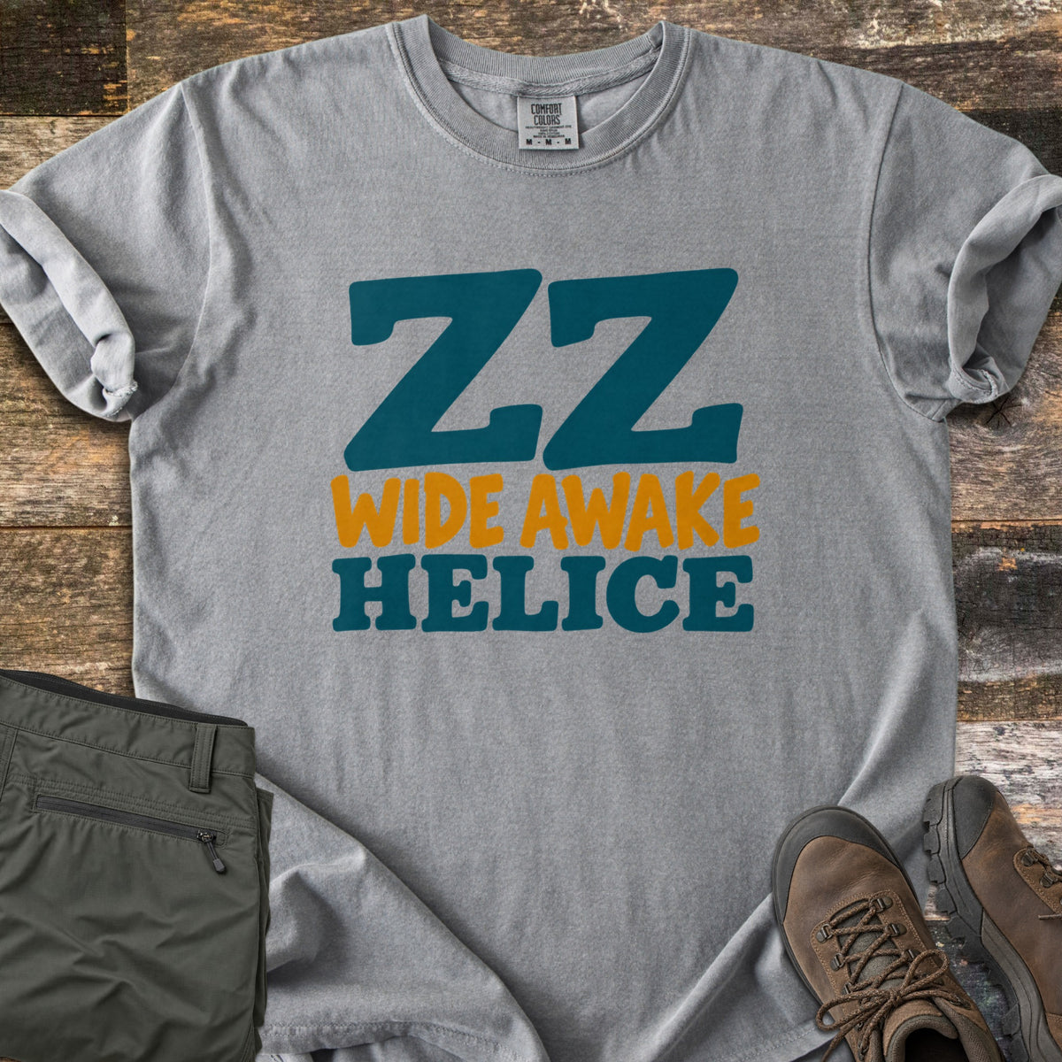 ZZ Wide Awake Comfort Colors T-shirt in Grey, laid flat. Large teal letters read ZZ, with Wide Awake in orange and Helice in teal below. The sleeves are rolled on a rustic wood floor beside olive cargo pants and brown leather hiking boots.