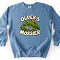 Older & Mossier Comfort Colors crewneck sweatshirt in Blue Jean, laid flat. A smiling cartoon rock covered in green moss and wearing a red polka-dot party hat sits between the words Older & Mossier in cream bubble letters. The sweatshirt lies on a white background.