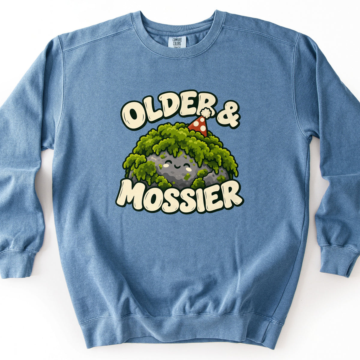 Older & Mossier Comfort Colors crewneck sweatshirt in Blue Jean, laid flat. A smiling cartoon rock covered in green moss and wearing a red polka-dot party hat sits between the words Older & Mossier in cream bubble letters. The sweatshirt lies on a white background.