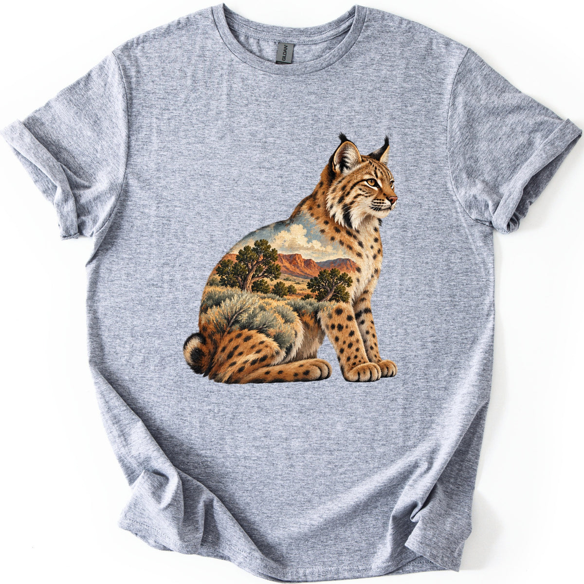Bobcat Sagebrush Canyon Comfort Colors T-shirt in Sport Grey, laid flat. A painted bobcat sits facing right, its body filled with a desert scene of red canyon cliffs, green trees, sagebrush and clouds. No words are printed. The shirt lies on a white surface with cuffed sleeves.