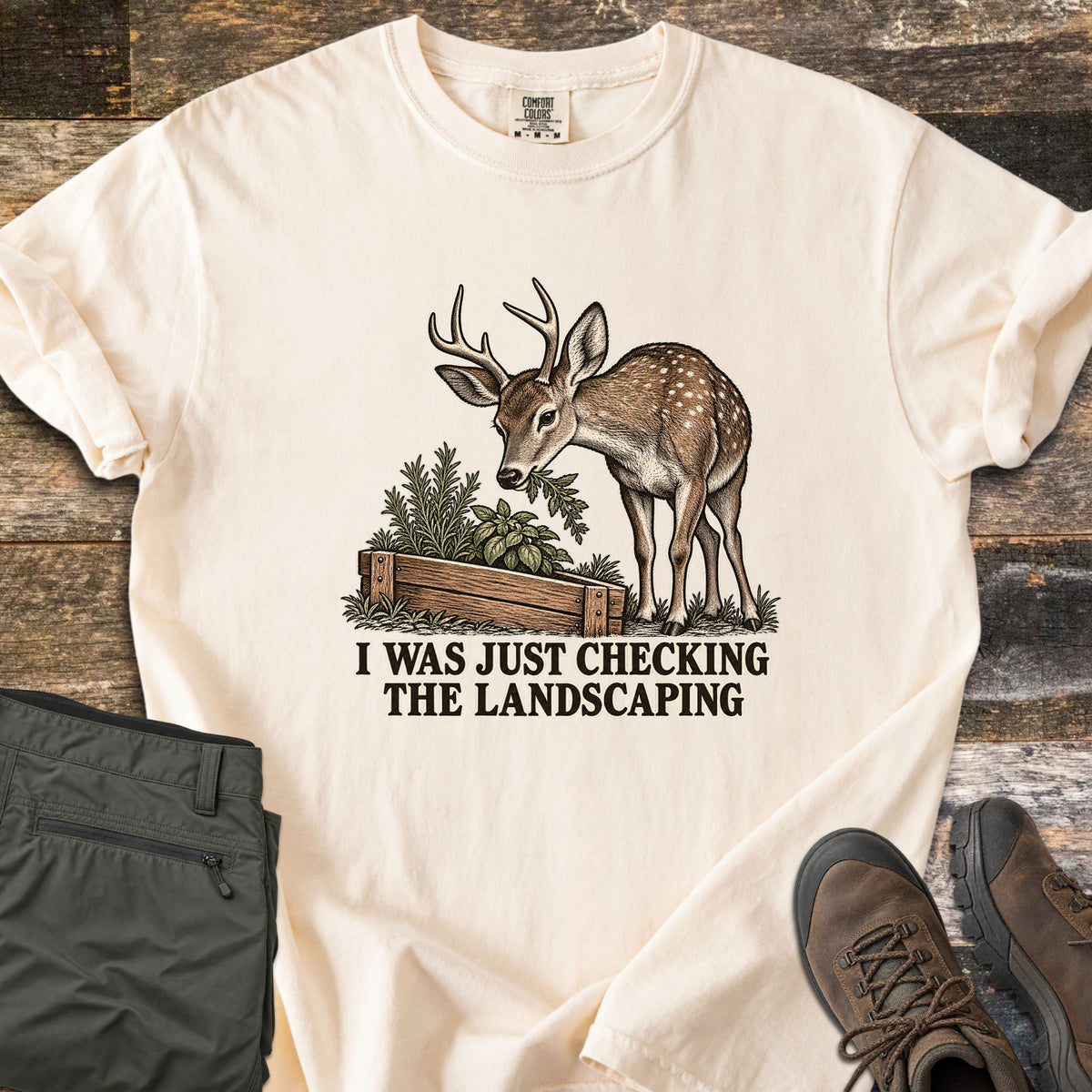 I Was Just Checking the Landscaping Comfort Colors T-shirt in Ivory, laid flat. A young antlered deer with white spots nibbles herbs from a wooden raised garden bed above the words I Was Just Checking the Landscaping. Green pants and brown hiking boots lie beside it on weathered wood.