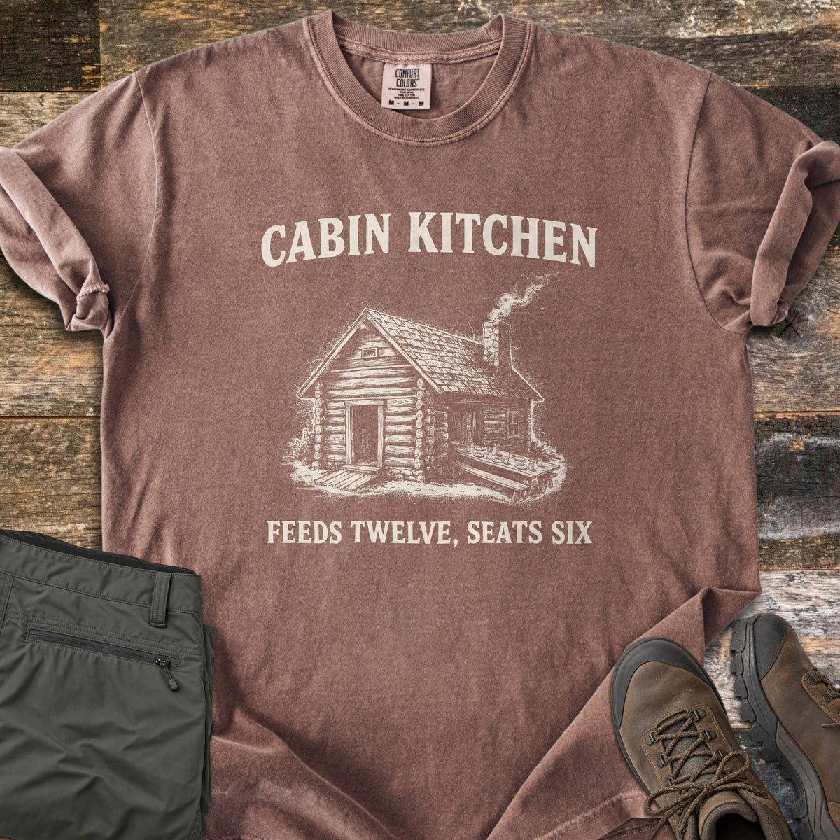 Cabin Kitchen Comfort Colors T-shirt in Espresso, laid flat. An off-white sketch of a smoking log cabin with a long outdoor table sits between the words Cabin Kitchen and Feeds Twelve, Seats Six. Olive hiking pants and brown hiking boots lie beside it on weathered wood.