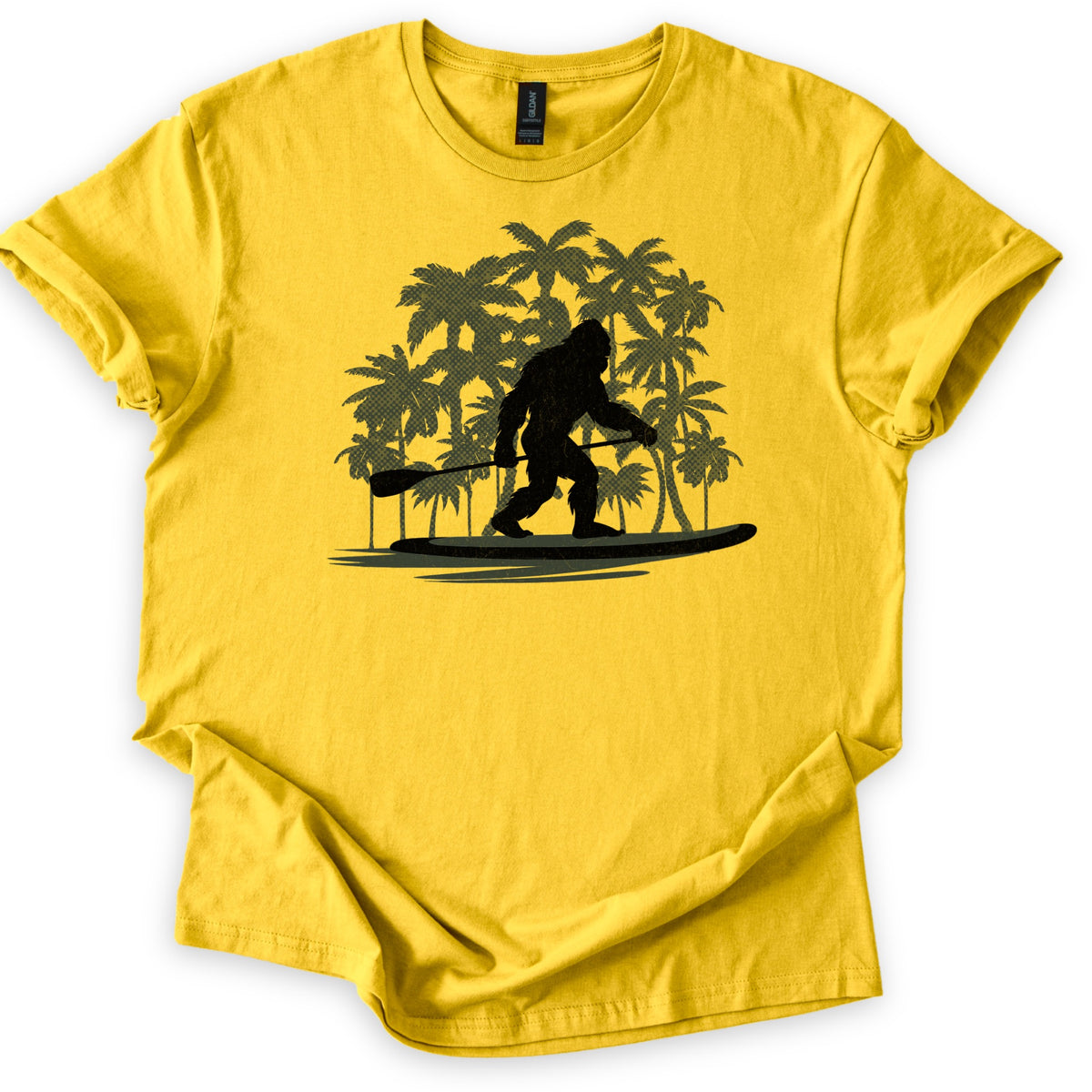 Paddle Board Squatch Comfort Colors T-shirt in Daisy, laid flat. A black silhouette of Bigfoot stands on a paddle board holding a paddle, in front of a cluster of olive green palm trees, with no words printed. The shirt lies on a white background with rolled sleeves.